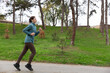 © Ekaterina - Athletic woman jogging on a pathway surrounded by lush green trees, wearing a blue jacket and brown leggings, showcasing a healthy lifestyle and outdoor fitness activity