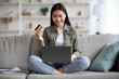 © Prostock-studio - Cheerful young asian woman sitting on couch in living room and using laptop and credit card, checking new webstore for online shopping or banking, home interior, copy space, catching sales