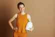 © Alliance - A smiling woman in orange tank top and leggings holds protein powder container, posing with hand on hip. Against soft brown background, this medium shot embodies active lifestyle and wellness