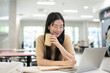 © bongkarn - Glasses asian woman college student holding pen and coffee while smiling looking at laptop on table.