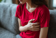 © © Ai Factory - Young woman in a red shirt sitting on a sofa and holding her chest in pain, heart attack or anxiety concept for healthcare and medical emergency at home