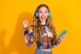 Young woman with blue headphones smiles while holding a phone against a bright yellow background showcasing casual fashion and music lifestyle
