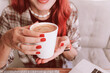 © EdNurg - Red haired woman with red manicure holding a warm white coffee mug by her laptop, relaxing in a cozy work from home moment