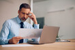 © Drazen - Pensive businessman analyzing reports while working on laptop in office.