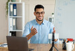 © Prostock-studio - Remote learning concept. Joyful Arab teacher giving online English lesson, using smartphone and laptop at home office. Young male tutor conducting web conference with students