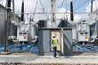 © JD Studio - Male electrical engineer inspecting control cabinet of large power transformers at high voltage substation to monitor energy distribution grid and industrial utility infrastructure.