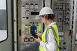 © JD Studio - Female electrical engineer operating protection relay and control system on indoor panel while holding digital tablet for maintenance audit at high voltage substation facility.