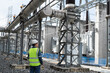 © JD Studio - Professional electrical engineer inspecting outdoor high voltage substation infrastructure and power transformers. Technician in safety vest monitoring energy grid equipment.