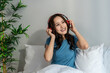 © Kawee - Asian woman listen to music from headphones while sitting on bed in house.