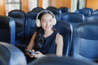 © Tj - Young Asian woman smiling with headphones, using smartphone while seated on public transit, enjoying music and a relaxed solo commute, browsing and staying connected