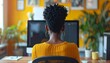 © Roberto - a black woman with short hair wearing a yellow blouse shirt working on computer sitting at desk in the office, seen from behind