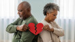 © Iryna - Elderly African-American couple holding torn heart halves facing away in home, relationship separation and mature breakup acceptance