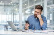 © Liubomir - Bored businessman feeling unproductive and procrastinating, sitting at office desk checking smartphone while laptop and work documents remain untouched, representing job burnout and distraction