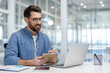 © Liubomir - Positive bearded businessman working on a laptop, smiling and writing in a notebook while participating in a virtual conference or remote work session in a modern office