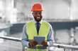 © Prostock-studio - A construction worker smiles while holding a tablet in an industrial setting. He wears a bright safety vest and hard hat. The worker appears engaged in project management tasks at the site.