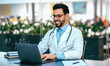 © Prostock-studio - A doctor is sitting at a desk in a modern office setting. He is smiling while using a laptop. There are plants in the background. Books are placed nearby.