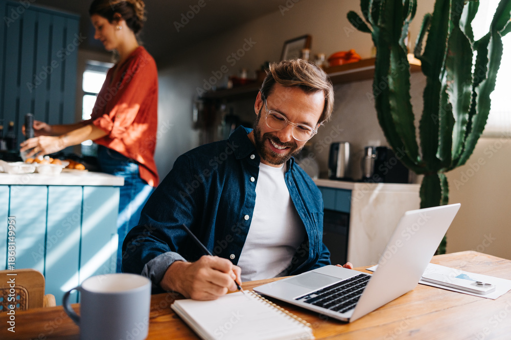Man at home working on laptop and writing in notebook