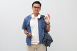 © ArvStd - Young asian male student with glasses smiling expressing emotion of success, showing okay gesture and holding digital tablet computer, wearing casual clothes, backpack and headphones around neck.