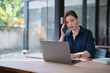 © Tj - Businesswoman multitasking, engaging in a phone call while simultaneously working on a laptop at a desk, demonstrating efficient remote work and communication