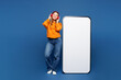 © ViDi Studio - Full body young happy woman in yellow sweater casual clothes big huge blank screen mobile cell phone smartphone with area listen music in headphones isolated on plain blue background studio portrait