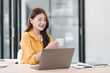 © PRIME STOCK LAB - A smiling young Asian woman working on a laptop while holding a coffee cup in a bright modern office.