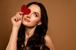 © Beauty Hero - Young brunette woman with heart-shaped accessory on beige background expressing beauty and charm