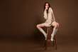 © Beauty Hero - Young woman in an elegant beige sweater posing gracefully while seated on a wooden stool