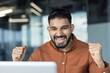 © Liubomir - Enthusiastic young man with a beard cheering and clenching fists while looking at a laptop screen, celebrating success or a win in a modern office environment