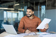© Liubomir - Young indian businessman smiling while reviewing documents and taking notes at a laptop-filled desk, conveying productivity, confidence and professional success in a modern office setting