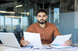 © Liubomir - Serious indian businessman sitting at an office desk, analyzing documents while a laptop and paperwork are spread around, representing professional work and diligent review of data