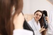 © New Africa - Smiling woman in bathrobe styling her hair with hairdryer and brush near mirror in bathroom