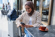 © Jovo Jovanovic/Stocksy - Woman in hijab smiles at phone with coffee nearby.