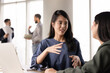 © fizkes - Positive young Asian professional woman talking to female colleague at workplace, speaking to business colleague at work table, explaining task, creative idea for teamwork