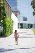 © Masakazu Tokashiki - A woman is walking down a sidewalk in front of a tall building. She is wearing a pink shirt and shorts