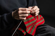 © ugis - Close-up of a person knitting with red and green yarn, creating a striped fabric. The individual is wearing dark clothing. No visible logos or faces
