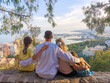 © Ina - Rear view of a family sitting together on a hilltop viewpoint, enjoying the panoramic view of Malaga city and the Mediterranean Sea, Spain, on a sunny day.