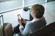 © Wavebreak Media - Sitting on metal bench in airport departure lounge, woman holding smartphone near spinner suitcase