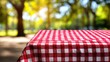 © mila103 - A checkered cloth covers a picnic table in an outdoor park setting. Sunlight filters through the trees while people enjoy their time in the background.
