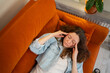 © wifesun - Young woman suffering from a headache or migraine, lying on an orange sofa, pressing her temples, eyes closed in discomfort, reflecting symptoms of illness, stress, and chronic pain at home