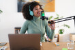 © Studio Marmellata - A woman with headphones speaks into a microphone while working on a laptop, creating content for her broadcast.