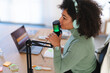 © Studio Marmellata - A woman with headphones speaks into a microphone while recording a podcast. A laptop is open on the desk beside her.