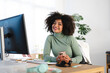 © Studio Marmellata - A smiling woman with curly hair sits at her desk, holding a smartphone. She is dressed in a green turtleneck and has a computer and headphones in front of her.
