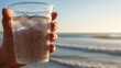 © MDT - Close-up view of hand holding plastic cup filled with clear sparkling water and ice cubes on sunny beach day