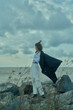 © SHOTPRIME STUDIO - Windy coastal scene with a flowing cape and white attire on a rocky shore, moody clouds above, capturing freedom, fashion and adventure in a dramatic ocean setting