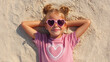 © Helena GARCIA - niña de 7 años tumbada en la arena de una playa con los brazos detras de la cabeza. Sonrie y lleva gafas de sol rosas conforma de corazon, coletas y camiseta rosa con un corazon blanco
