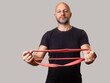 © mark_gusev - Athletic man in his 40s or 50s holding red resistance band. Male model wearing black shirt with slim body type and on grey background with popular sport equipment. Sport and fitness concept.