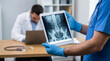© DebbyMelindaYanti - A medical professional in blue scrubs and gloves reviews a human spine and pelvis X-ray on a digital tablet, demonstrating modern medical diagnostics