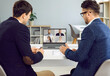 © Studio Romantic - Two businessmen sitting at office desk with laptop computer, using modern video conferencing app, having online meeting with remote team of business people, and discussing work projects together