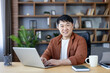 © Liubomir - Smiling asian man in smart-casual attire types on a laptop at a wooden desk in a modern home office, looking at the camera with confident, relaxed professionalism
