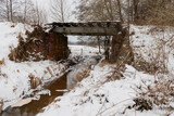 Strumień płynący pod starym mostem kolejowym. W poprzek strumienia jest tama z gałęzi zbudowana przez bobry. Jest zima, brzegi i tamę pokrywa warstwa śniegu.